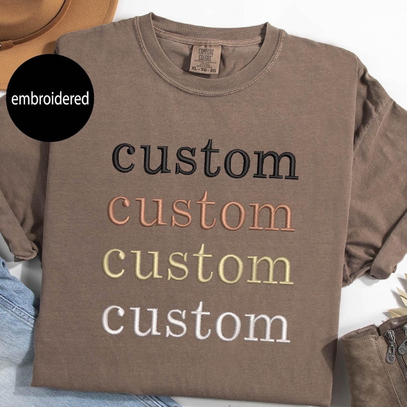 May include: A brown t-shirt with the word "custom" embroidered on it in four different colors: black, peach, yellow, and white.