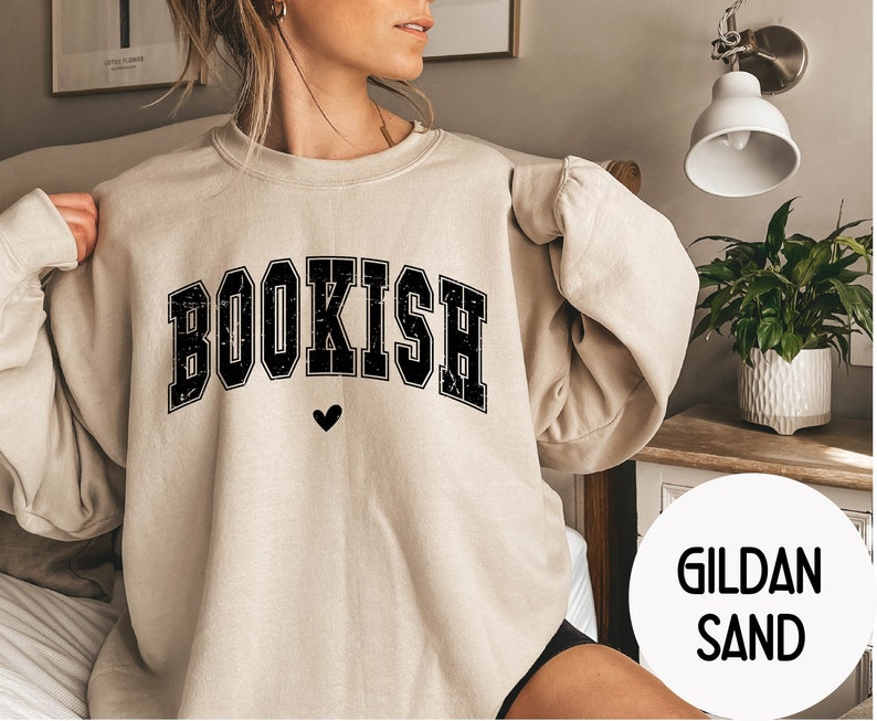 May include: A beige sweatshirt with the word "BOOKISH" printed in black with a small heart below it. The sweatshirt is made by Gildan and is in the color Sand.