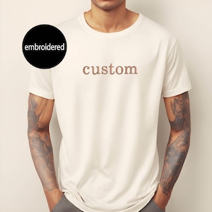May include: White t-shirt with the word "custom" embroidered in brown on the front.