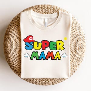 May include: A white t-shirt with a colorful graphic of a Super Mario character wearing a hat and the text "Super Mama".