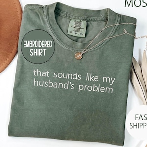 May include: An olive green embroidered shirt with the text "that sounds like my husband's problem" in white. A gold necklace with a pendant is on the shirt. The shirt has a Comfort Colors tag.