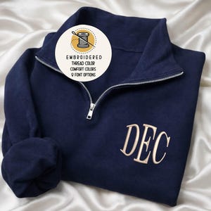May include: Navy blue quarter-zip sweatshirt with embroidered initials "DEC" in a light tan color. The sweatshirt has a silver zipper and a round graphic with text that reads "Embroidered Thread Color, Comfort Colors, 9 Font Options."