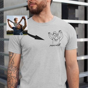 May include: A light gray t-shirt featuring a line art illustration of a father and child, with the text "papa + lill" below the graphic. A small photo of a father and child is on the upper left. The shirt is worn by a person with a beard.