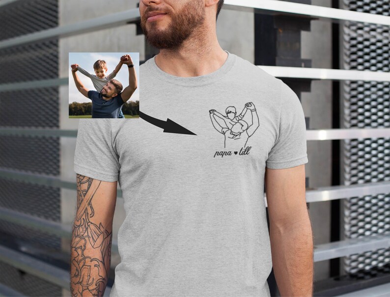 May include: A light gray t-shirt featuring a line art illustration of a father and child, with the text "papa + lill" below the graphic. A small photo of a father and child is on the upper left. The shirt is worn by a person with a beard.