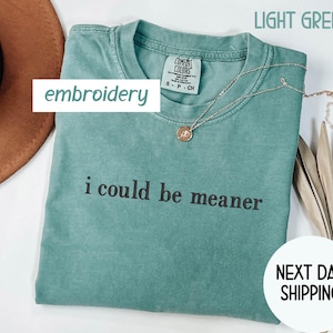 May include: Light green t-shirt with the phrase "i could be meaner" in black text. The shirt has a crew neck and a small tag. A gold necklace with a pendant is draped across the shirt. The words "embroidery" and "next day shipping" are also visible.