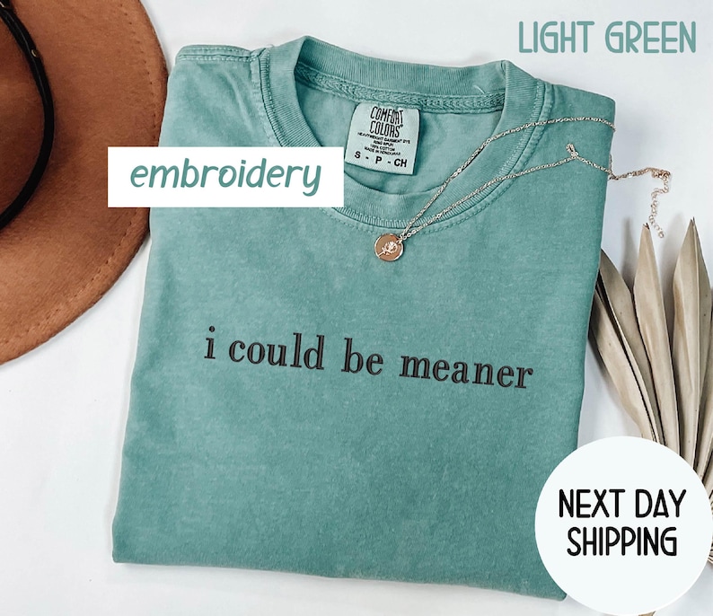 May include: Light green t-shirt with the phrase "i could be meaner" in black text. The shirt has a crew neck and a small tag. A gold necklace with a pendant is draped across the shirt. The words "embroidery" and "next day shipping" are also visible.