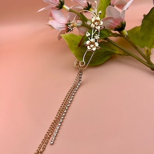 Puede incluir: Pendiente de oreja dorado con diseño floral y una cadena colgante con pedrería. El pendiente de oreja está hecho de metal y tiene un aspecto delicado y femenino.