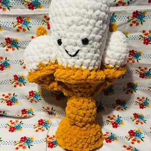 May include: A crocheted ice cream cone with three scoops of white ice cream and a smiling face. The ice cream cone is sitting on a yellow crocheted stand.