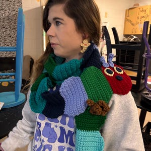 May include: A colorful, handmade crochet scarf in shades of green, blue, and red, designed to resemble a caterpillar. The scarf is worn around the neck. The person is wearing a gray sweatshirt with the text "HIGH" and "CHILDREN".