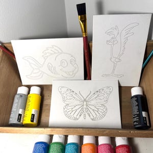 May include: Three paint-by-number canvases with outlines of a fish, a roadrunner, and a butterfly. The canvases are in a wooden box with paint tubes in front of them. The paint tubes are in various colors, including gray, yellow, blue, green, orange, and red.