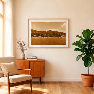 Puede incluir: Una impresión de paisaje enmarcada que muestra una escena costera en tonos sepia con montañas, agua y pequeñas embarcaciones, exhibida en una pared clara sobre un mueble de madera, un sillón beige y una planta de higuera de hoja.