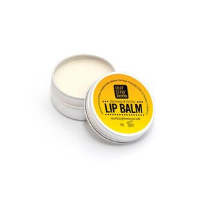 May include: A silver tin of lip balm with a yellow and black label. The label states "Lip Balm" and "Beeswax & Honey." The lip balm is a pale yellow colour. The product is from ourtinybees.co.uk.