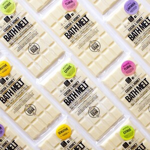 May include: Multiple bath melts in clear packaging, arranged diagonally. Each bar is white with a grid pattern and labeled "Cocoa & Honey BATH MELT." Round stickers indicate scents like "Citrus," "Floral," and "Lavender."