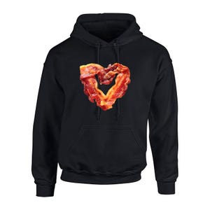 May include: Black hoodie featuring a heart formed from cooked bacon. The bacon is a mix of red and brown, contrasting against the black fabric. The hoodie has a front pocket and a drawstring hood, typical of this garment.
