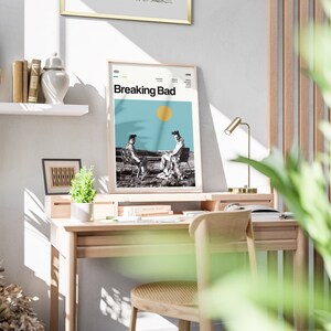 May include: A framed poster with the title "Breaking Bad" in white text on a blue background. The poster features a black and white image of two people sitting on chairs in a desert landscape. The poster is hanging on a white wall above a wooden desk.