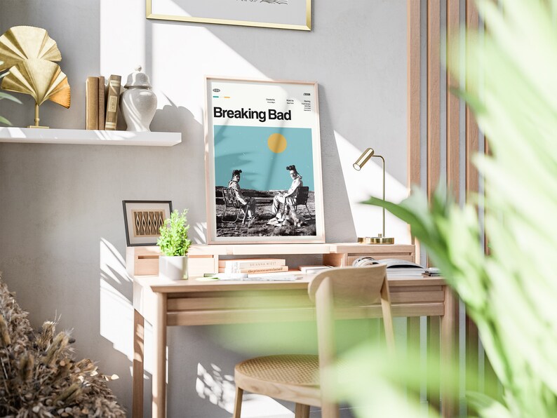 May include: A framed poster with the title "Breaking Bad" in white text on a blue background. The poster features a black and white image of two people sitting on chairs in a desert landscape. The poster is hanging on a white wall above a wooden desk.