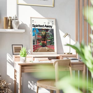 Może przedstawiać: Oprawiony plakat z napisem "Spirited Away" przedstawia żywą scenę z czerwonym mostem i tradycyjną japońską architekturą. Plakat jest umieszczony na drewnianym biurku z małą lampą i krzesłem. Widoczna jest r&oacute;wnież mała roślina doniczkowa i książki.