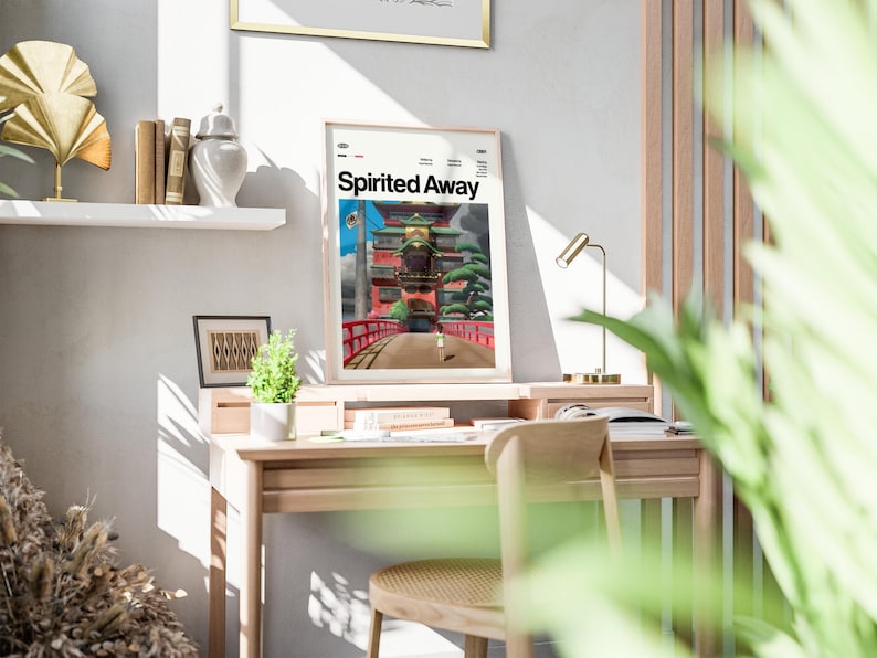 Może przedstawiać: Oprawiony plakat z napisem "Spirited Away" przedstawia żywą scenę z czerwonym mostem i tradycyjną japońską architekturą. Plakat jest umieszczony na drewnianym biurku z małą lampą i krzesłem. Widoczna jest r&oacute;wnież mała roślina doniczkowa i książki.