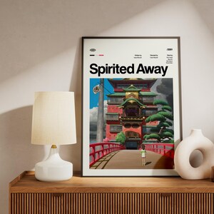 Może przedstawiać: Oprawiony plakat z tytułem "Spirited Away" w kolorze białym. Grafika przedstawia kolorowy japoński budynek z czerwonym mostem, postacią i zielonymi drzewami. Plakat jest eksponowany na drewnianej powierzchni, obok lampy i dekoracyjnych wazon&oacute;w.