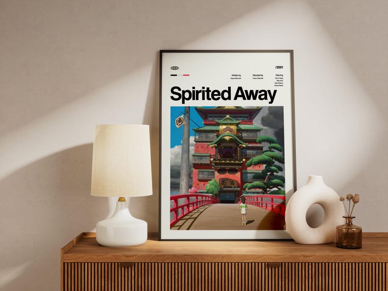 Może przedstawiać: Oprawiony plakat z tytułem "Spirited Away" w kolorze białym. Grafika przedstawia kolorowy japoński budynek z czerwonym mostem, postacią i zielonymi drzewami. Plakat jest eksponowany na drewnianej powierzchni, obok lampy i dekoracyjnych wazon&oacute;w.
