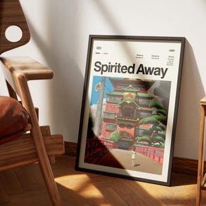 Może przedstawiać: Oprawiony plakat z tytułem "Spirited Away" w kolorze białym. Grafika przedstawia kolorową scenę z czerwonym mostem, dużym budynkiem i postacią odchodzącą. Plakat jest w czarnej ramie i opiera się o białą ścianę.