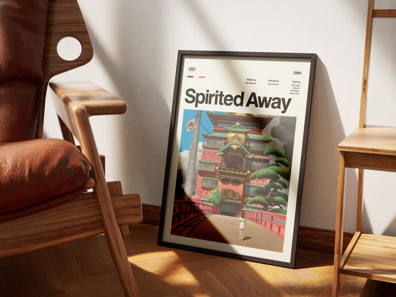 Może przedstawiać: Oprawiony plakat z tytułem "Spirited Away" w kolorze białym. Grafika przedstawia kolorową scenę z czerwonym mostem, dużym budynkiem i postacią odchodzącą. Plakat jest w czarnej ramie i opiera się o białą ścianę.