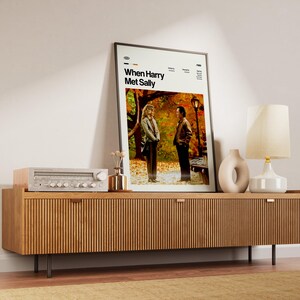 May include: A framed movie poster for "When Harry Met Sally" stands on a wooden cabinet. The cabinet has a ribbed design and holds a stereo, a vase, and a lamp. The poster features two people in a park setting.