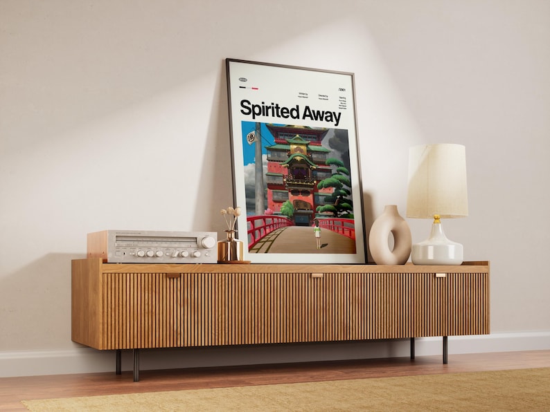 Może przedstawiać: Oprawiony plakat z napisem "Spirited Away" jest wyświetlany na drewnianej szafce. Plakat przedstawia kolorową ilustrację budynku z czerwonym mostem. Na szafce znajduje się r&oacute;wnież zabytkowy zestaw stereo, wazon i lampa.