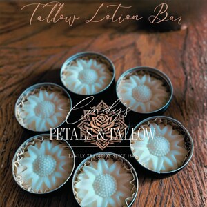 Peut inclure: Six barres de lotion au suif blanches en forme de fleurs, chacune dans une petite boîte ronde. Les boîtes sont disposées en cercle sur une surface en bois. Le texte "Tallow Lotion Bar" et "Cindy's Petals & Tallow" sont visibles.