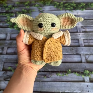 Op de afbeelding: Een gehaakt groen en bruin pluche speelgoed in de vorm van een personage uit de Star Wars-franchise. Het speelgoed draagt een bruin gehaakt jasje met witte afwerking.