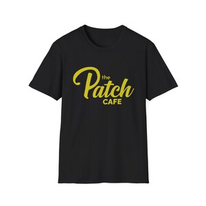 Puede incluir: Camiseta negra con el texto "the Patch Cafe" en amarillo, con una fuente cursiva. La camiseta es de cuello redondo, adecuada para uso casual. El texto está centrado en el pecho.