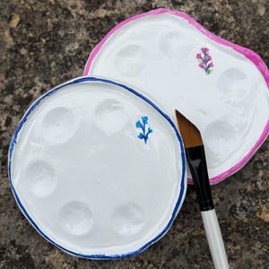 May include: Two white paint palettes with floral accents and a paintbrush. One palette has a blue border and a blue floral design, the other has a pink border and a pink floral design. The paintbrush has a white handle and brown bristles.