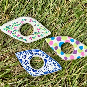 May include: Three decorative, eye-shaped ceramic pieces with a central hole, resting on grass. One is white with pink and green floral designs, another is blue with white floral patterns, and the third is green with purple, blue, and pink polka dots.