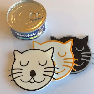 May include: A can of Tastefuls flaked tuna cat food next to three cat-shaped coasters. The coasters are white, orange, and black with closed eyes and smiling faces. The coasters are made of a glossy material.