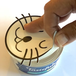 May include: A white plastic jar with a black and white cartoon cat lid. The lid is being lifted off the jar. The jar label reads "Tastefuls Tuna Entree in Gravy".