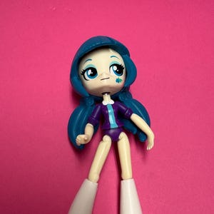 Puede incluir: Una pequeña muñeca de plástico con cabello azul, ojos grandes azules y una pequeña marca de flor azul en la mejilla izquierda. La muñeca viste una chaqueta morada con una franja central azul claro, parte inferior morada y botas blancas.