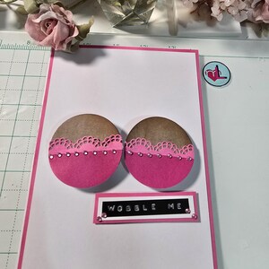 May include: A pink and brown card with two pink and brown circles with white lace trim and pink rhinestones. The card has a black banner with the text "WOBBLE ME" in white letters and pink rhinestones.