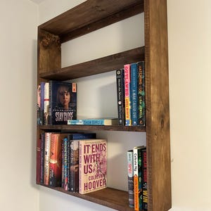 May include: A dark stained wooden bookshelf with three shelves, mounted on a white wall. The shelves hold a variety of books with colorful spines. The bookshelf is rectangular and has a simple, modern design.