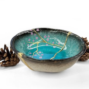 May include: A ceramic bowl with a teal interior and a dark rim. The bowl features gold-colored kintsugi repairs and is decorated with pink cherry blossoms. Two pine cones are on either side of the bowl.