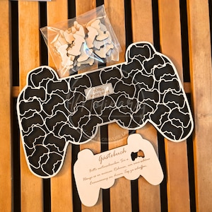 Peut inclure: Un livre d'or en bois noir et blanc en forme de manette de jeu vidéo, avec un petit panneau en forme de manette. Le livre d'or présente un motif découpé de formes de manettes. Un petit sac de découpes de manettes en bois est également présent. Le texte "JÖRG 20.09.2025" est visible.