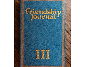 May include: A blue journal with the words "friendship Journal" in gold lettering. The journal has a gold border and the Roman numeral "III" in gold. The journal is on a wooden surface.