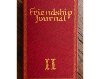 May include: A red leather-bound journal with the words "Friendship Journal" in gold lettering. The journal has a gold border and the Roman numeral "II" in gold at the bottom. The journal is on a wooden surface.