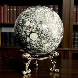 May include: A large, spherical decorative object with a black and white patterned surface, resting on an ornate silver-colored stand. The sphere's design features a mix of dark and light mottled patterns. The stand has a detailed, scrolled design.