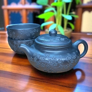 May include: A dark gray ceramic teapot with a matching lid and two teacups. The teapot and cups feature a floral pattern. The set is displayed on a wooden surface, with a blurred background of plants and furniture.