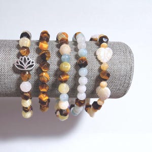 May include: Five beaded bracelets displayed on a gray fabric roll. The bracelets feature a variety of gemstones, including tiger's eye, white, and light blue beads. One bracelet has a silver lotus charm. The bracelets are a mix of earth tones and pastel colors.