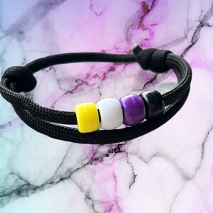 Könnte beinhalten: Ein schwarzes Armband aus Kordel mit gelben, weißen und violetten Perlen. Das Armband liegt auf einem Marmoruntergrund.