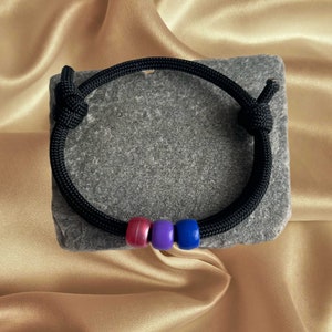 May include: A black paracord bracelet with three beads in the colours of the bisexual pride flag: pink, purple and blue.