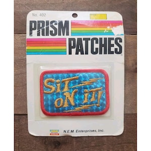 Op de afbeelding: Een rechthoekige geborduurde patch op een witte achtergrondkaart met het label Prism Patches No. 402. De patch heeft een blauwe holografische achtergrond met een rode rand en goudkleurige geborduurde tekst Sit on it! met bliksemschichten.