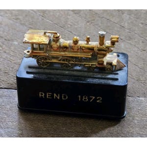 May include: A miniature gold-toned metal steam locomotive train model mounted on a rectangular black base. The base features the text REND 1872 printed in gold lettering on the front side. The train rests on two parallel tracks.