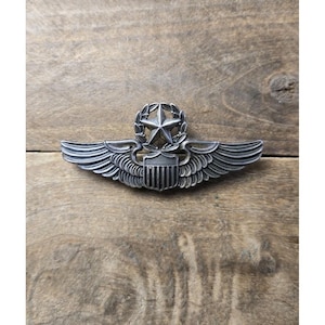 Pode incluir: Um emblema de metal em tom prateado de estilo militar com um escudo central com listras verticais, encimado por uma estrela de cinco pontas rodeada por uma coroa circular, com asas emplumadas estilizadas que se estendem horizontalmente.